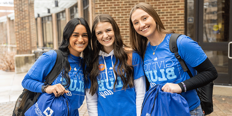 Three smiling female students stand outside the Hulman Memorial Student Union. All three wear Give to Blue Day t-shirts. All three have long, dark hair. The one on the right and on the left are holding blue drawstring book bags.